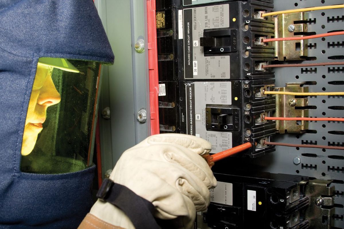 Technician in an arc-rated hood with a tinted face shield and leather-protected gloves testing a moulded-case circuit breaker inside a live panel