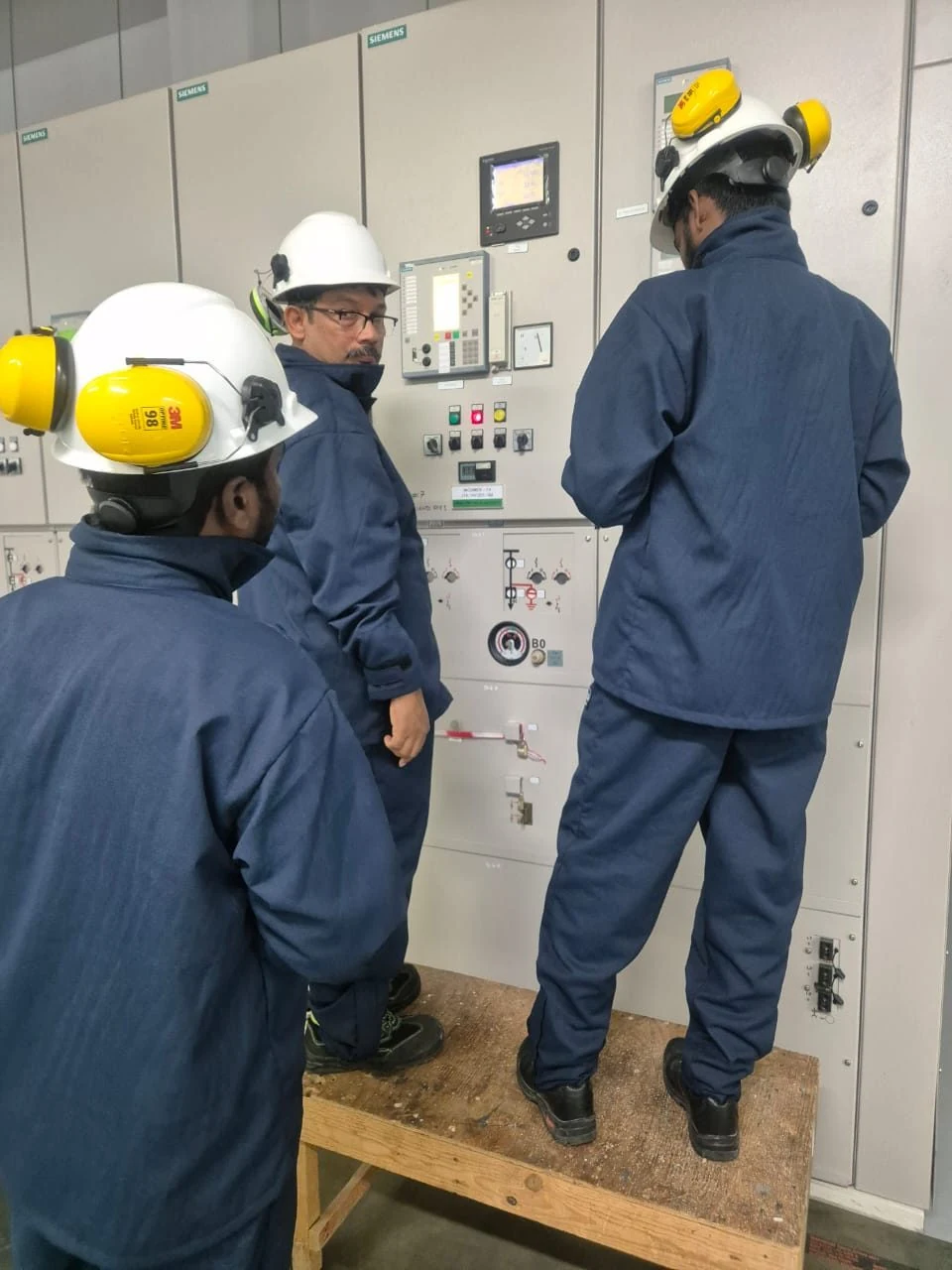 Three VB engineers in blue arc rated coveralls and 3M Peltor hard hats reading a Siemens MV switchgear relay HMI during the VB Arc360 ASSESS walkdown · capturing relay protection settings on the spot · real field photo