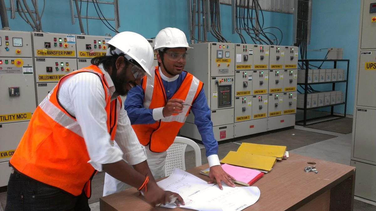 VB engineer and client engineer reviewing the plant single line diagram on a table inside the UTILITY MCC room · CAPACITOR BANK and APFC PANEL cells visible on the labeled MCC behind them · this is the VB Arc360 ASSESS on-site data collection walkdown · real field photo