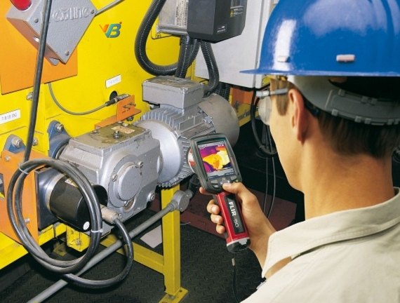 Engineer in a hard hat scanning a running motor and its terminal box with a thermal imager